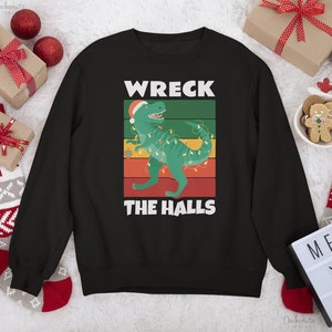 Puede incluir: Sudadera negra con un diseño de un dinosaurio verde con gorro de Papá Noel y luces navideñas. El texto "WRECK THE HALLS" está presente. Ropa festiva navideña.