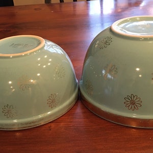 May include: Two turquoise ceramic mixing bowls with gold daisy patterns. The larger bowl has a gold rim.