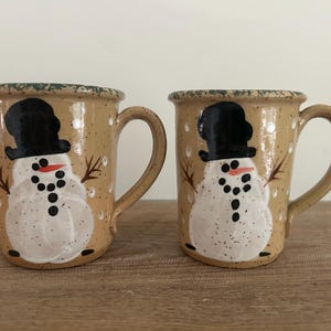 May include: Two ceramic mugs with a snowman design. The mugs are tan with a speckled finish and feature a hand-painted snowman with a black top hat, coal eyes, and a carrot nose. The mugs have brown handles and are decorated with white dots.
