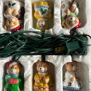 May include: A set of six decorative teddy bear light covers arranged in a white styrofoam tray with a green electrical cord. Each bear features distinct clothing, including hats, scarves, and dresses in colors like red, green, yellow, and white.
