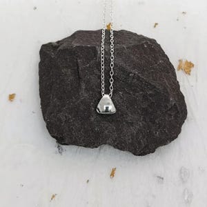 May include: A silver necklace featuring a triangular pendant. The pendant, with a smooth, reflective surface, hangs from a fine chain. The necklace is presented on a dark grey rock against a white backdrop.