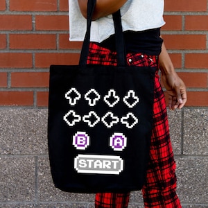 May include: Black tote bag featuring a pixelated design of directional arrows, the letters "B" and "A" in purple boxes, and the word "START" in a gray box. The bag is held against a brick wall.