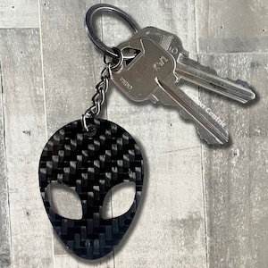 May include: Black and gray alien-shaped keychain with a carbon fiber weave pattern. The keychain is attached to a silver key ring and chain, with two silver keys. The alien has large, oval eyes and a small mouth.