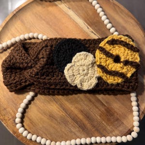 May include: A brown crocheted headband with three decorative elements: a black circle, a beige flower, and a yellow and brown striped donut. The headband is displayed on a wooden tray with a beaded garland.
