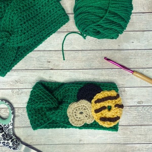 May include: Green crocheted headband with a brown, yellow, and white crocheted flower design. The headband is on a wooden surface with a ball of green yarn, a pair of scissors, and a crochet hook.