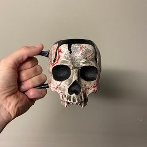 Puede incluir: Una taza de cerámica blanca en forma de cráneo con detalles de pintura negra y roja. La taza tiene un asa negra y un efecto de pintura negra que gotea en la parte superior.