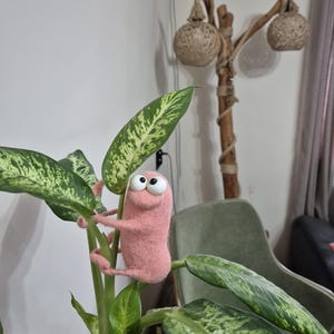 May include: A pink felted creature with large googly eyes clings to a green and white patterned plant. The whimsical figure has small arms and legs, adding a playful touch to the indoor scene. The plant's leaves are large and oval-shaped.