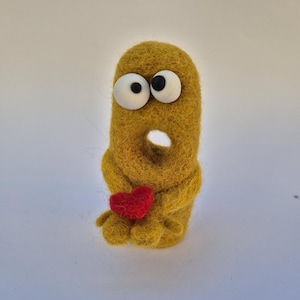 May include: A handmade, whimsical felt creature in a mustard yellow color, with large googly eyes and a red heart. The creature has a hole in its body and small feet, giving it a unique and playful appearance.