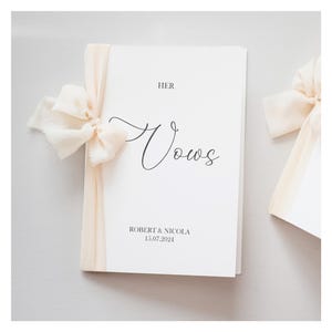 Op de afbeelding: Een wit boekje met het woord "Vows" in elegant schrift, vastgebonden met een crèmekleurig lint. Het woord "HER" staat boven het woord "Vows" gedrukt. De namen "ROBERT & NICOLA" en de datum "15.07.2024" staan onderaan gedrukt.
