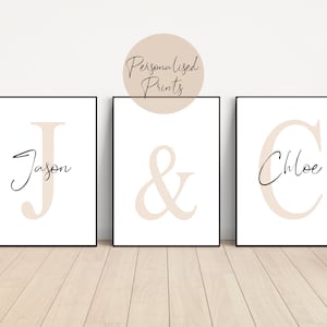 May include: Three white framed prints with beige letters. The first print has a large beige letter J with the name Jason written in cursive below it. The second print has a large beige ampersand symbol. The third print has a large beige letter C with the name Chloe written in cursive below it.