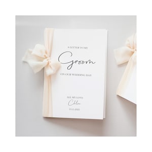 May include: A white wedding card with the text "A Letter to My Groom on Our Wedding Day" in elegant script. The card is tied with a cream-colored ribbon and bow. The name "Chloe" and the date "19.11.2023" are also printed on the card.