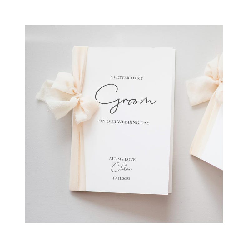 May include: A white wedding card with the text "A Letter to My Groom on Our Wedding Day" in elegant script. The card is tied with a cream-colored ribbon and bow. The name "Chloe" and the date "19.11.2023" are also printed on the card.