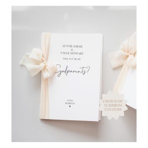 Puede incluir: Una tarjeta rectangular blanca con el texto impreso en negro AUNTIE SARAH & UNCLE STEWART WILL YOU BE MY Godparents? LOVE ISABELLE. La tarjeta está adornada con una cinta de tela color crema atada en un lazo en el lado izquierdo. Un gráfico en forma de estrella muestra el texto CHOICE OF 15 RIBBON COLOURS.