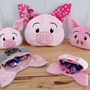 Puede incluir: Tres cestas de Pascua de peluche en forma de cerdo rosa con orejas caídas. Las cestas están abiertas y llenas de huevos de chocolate envueltos en papel de aluminio de colores.