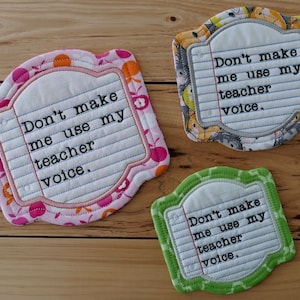 Op de afbeelding: Drie stoffen onderzetters met een roze, grijze en groene achtergrond. Elke onderzetter heeft een wit label met de tekst "Maak me niet mijn leraren stem gebruiken."
