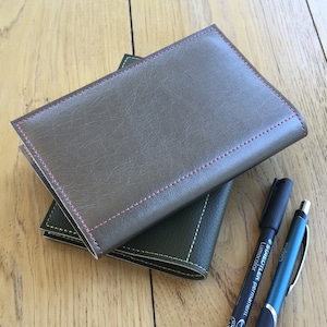 Puede incluir: Dos cuadernos encuadernados en cuero con costuras verdes, apilados sobre una superficie de madera. Un marcador negro y un bolígrafo azul están junto a los cuadernos.