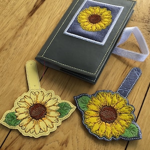 May include: Three leather bookmarks featuring embroidered sunflowers. The bookmarks are in shades of yellow, green, and blue. The bookmarks are displayed on a wooden surface.