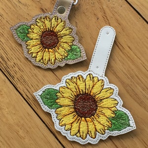 May include: Two white leather keychains with embroidered sunflowers. The sunflowers are yellow with brown centers and green leaves. One keychain has a metal ring for attaching keys.