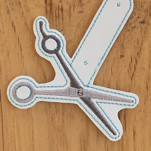 Puede incluir: Un parche de tela blanco con un diseño bordado de unas tijeras grises con costuras azules. Las tijeras están en posición cruzada.