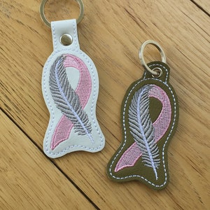 May include: Two keychains with pink and gray ribbon designs. The ribbon is embroidered with a feather design. One keychain is white and the other is green.
