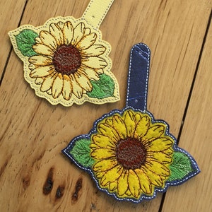 Puede incluir: Dos llaveros bordados con forma de girasol, uno con fondo amarillo y otro con fondo azul. Ambos llaveros presentan un diseño de girasol detallado con hojas verdes.