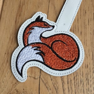 May include: Embroidered patch featuring a curled-up fox in shades of orange, white, and black. The fox is outlined in black stitching and sits on a white background with a rectangular extension. The patch is designed for sewing or ironing onto fabric.