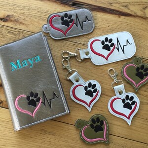 Può includere: Un portapassaporto in pelle argentata con il nome "Maya" ricamato sul davanti. Il portapassaporto presenta un cuore rosa con un'impronta di zampa nera e una linea di battito cardiaco. Ci sono cinque portachiavi con un design simile, che presentano un cuore rosso con un'impronta di zampa nera e una linea di battito cardiaco su uno sfondo bianco, grigio o verde.