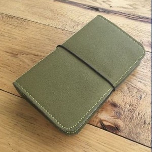 May include: A green leather notebook cover with a black elastic closure. The cover has a subtle texture and is stitched with yellow thread.
