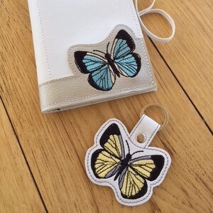 May include: A white leather notebook with the name "DIANE" embroidered on the front. The notebook has a butterfly design with blue and white wings. A matching butterfly keychain is also included.