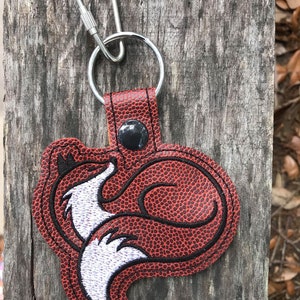K&ouml;nnte beinhalten: Ein rot-schwarzer, fuchsf&ouml;rmiger Schl&uuml;sselanh&auml;nger mit einem silbernen Schl&uuml;sselring und Karabinerverschluss. Das Fuchsmotiv hat wei&szlig;e und schwarze Stickdetails. Der Schl&uuml;sselanh&auml;nger besteht aus einem strukturierten Material und ist auf einer Holzoberfl&auml;che befestigt.