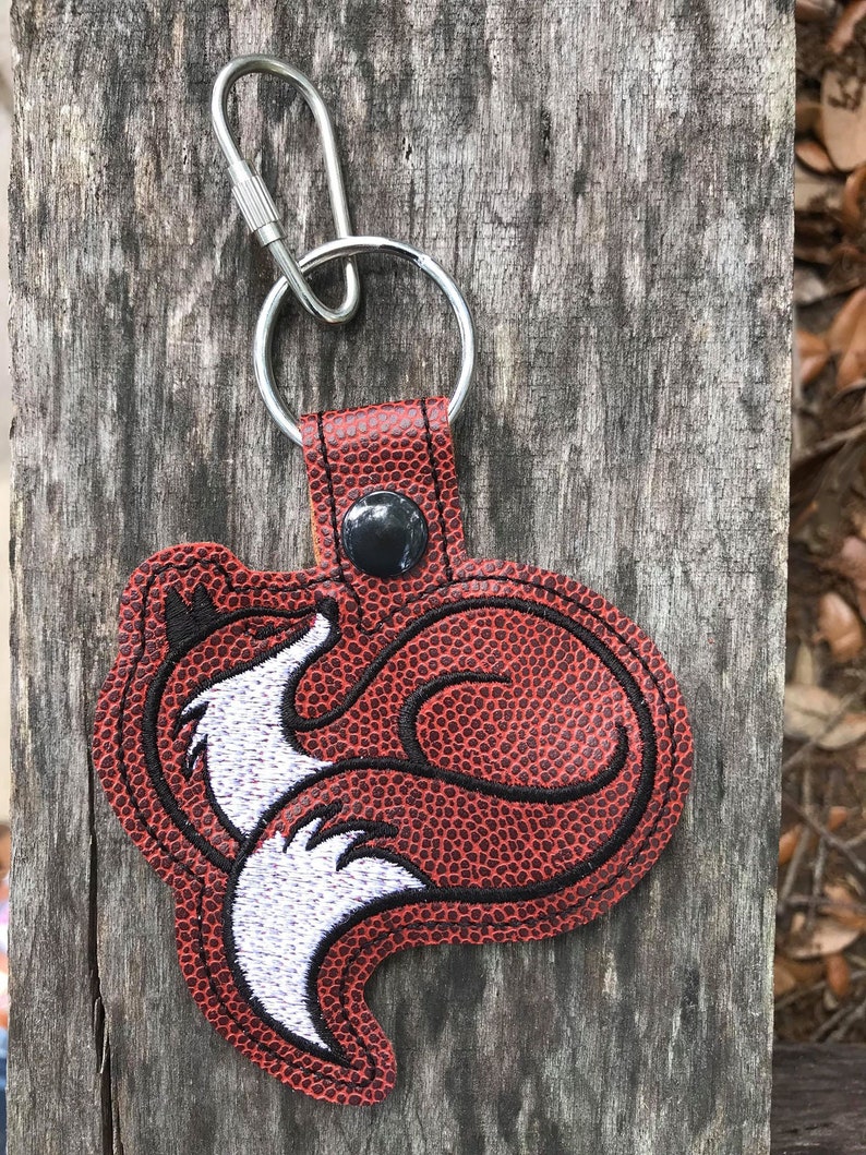 K&ouml;nnte beinhalten: Ein rot-schwarzer, fuchsf&ouml;rmiger Schl&uuml;sselanh&auml;nger mit einem silbernen Schl&uuml;sselring und Karabinerverschluss. Das Fuchsmotiv hat wei&szlig;e und schwarze Stickdetails. Der Schl&uuml;sselanh&auml;nger besteht aus einem strukturierten Material und ist auf einer Holzoberfl&auml;che befestigt.
