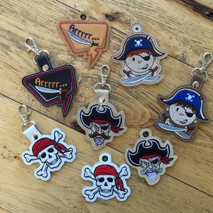 May include: Seven embroidered keychains with pirate themes.  The keychains feature a skull and crossbones, a pirate with an eyepatch, and a speech bubble with the text "Arrrr..."