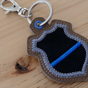 K&ouml;nnte beinhalten: Ein brauner Leder-Schl&uuml;sselanh&auml;nger mit einem blauen und schwarzen gestickten Schild. Das Schild hat eine d&uuml;nne blaue Linie dar&uuml;ber.