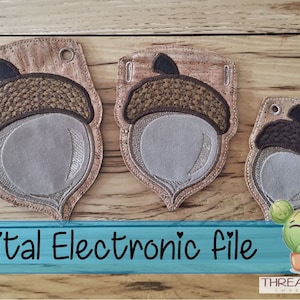 May include: Three acorn-shaped embroidery designs on a cork background. The designs are in various sizes and feature brown, tan, and grey stitching.