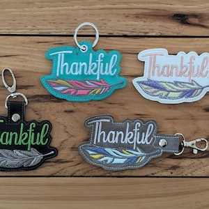 Puede incluir: Cuatro llaveros con un diseño de pluma y la palabra "Thankful" bordada en verde. Los llaveros son de diferentes colores: turquesa, blanco, negro y gris.