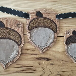 Puede incluir: Tres adornos en forma de bellota con respaldo de corcho y bordados marrones y beige. Los adornos est&aacute;n ensartados con una cinta negra.