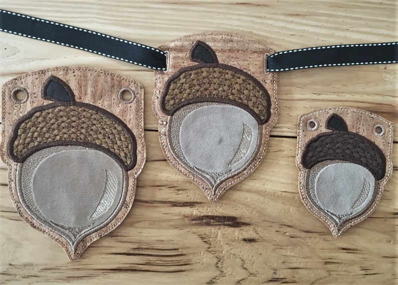 Puede incluir: Tres adornos en forma de bellota con respaldo de corcho y bordados marrones y beige. Los adornos est&aacute;n ensartados con una cinta negra.