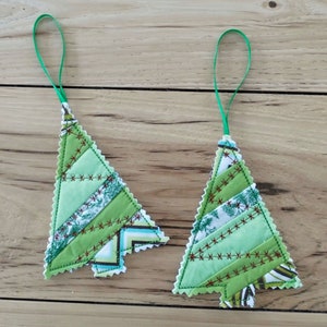 May include: Two handmade Christmas tree ornaments. Each tree is crafted from various shades of green fabric, stitched together with brown thread. They have a scalloped edge and a green ribbon loop for hanging. The ornaments are displayed on a wooden surface.