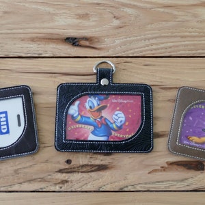 May include: Three black, brown, and gray leather ID badge holders with a clear plastic window. The holders have a stitched design around the edges. The left holder has a white card with the text "Prox Card II" and "HID". The middle holder has a colorful image of Donald Duck. The right holder has a colorful image of Pluto.