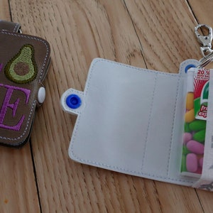 May include: Two small zippered pouches, one brown with the word "LOVE" embroidered in purple and an avocado design, and the other white with a clear window showing colorful candies.