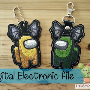 May include: Two embroidered keychains featuring characters from the video game Among Us. The characters are wearing bat wings and are in yellow and green colors. The keychains are on a wooden surface. Digital Electronic File.
