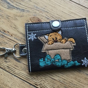 Puede incluir: Un llavero de cuero marrón con una bolsa con cremallera que presenta un diseño bordado de un gato en una caja con un lazo azul y copos de nieve.