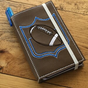 Op de afbeelding: Een bruin leren notitieboekje met een witte elastische band sluiting. De voorkant heeft een geborduurd ontwerp van een voetbal in een blauw en wit frame.