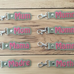 Op de afbeelding: Acht sleutelhangers met verschillende namen voor moeders, waaronder "Mum", "Mummy", "Mom", "Mommy", "Mama", "Mamma", "Madre" en "Mother". De sleutelhangers zijn gemaakt van groene stof met roze geborduurde letters.