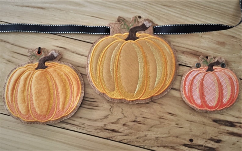 K&ouml;nnte beinhalten: Ein Herbstbanner mit drei gestickten K&uuml;rbissen. Die K&uuml;rbisse sind orange und gelb mit einem braunen Stiel und gr&uuml;nen Bl&auml;ttern. Das Banner besteht aus einem schwarzen Band mit wei&szlig;en Punkten.