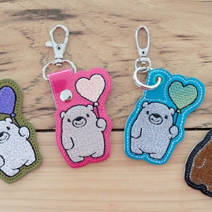 May include: Four keychains featuring embroidered animals holding heart-shaped balloons. The keychains are made of different colored leather and feature a silver metal ring and clasp. The animals include a white bear with a purple balloon, a gray bear with a pink balloon, a gray bear with a green balloon, and a brown bear with a green balloon.