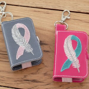 Puede incluir: Dos pequeños llaveros de cuero gris y rosa con un diseño de pluma blanca y cinta.