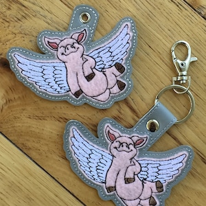 May include: Two grey felt keychains featuring a pink pig with wings. The pig is smiling and has its wings outstretched. The keychain on the right has a silver metal keyring.