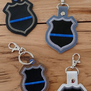 K&ouml;nnte beinhalten: Vier Schl&uuml;sselanh&auml;nger mit einem blauen Linien-Polizeibadge-Design. Die Schl&uuml;sselanh&auml;nger sind aus verschiedenen Materialien gefertigt, darunter Leder, Stoff und Metall. Jeder Schl&uuml;sselanh&auml;nger hat einen silbernen Metallschl&uuml;sselring.