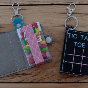 May include: Two small fabric pouches with keychains. The pouch on the left is gray with a blue trim and has a clear plastic window with a pack of Tic Tacs inside. The pouch on the right is black with a pink tic-tac-toe board and the text "Tic Tac Toe" in blue.